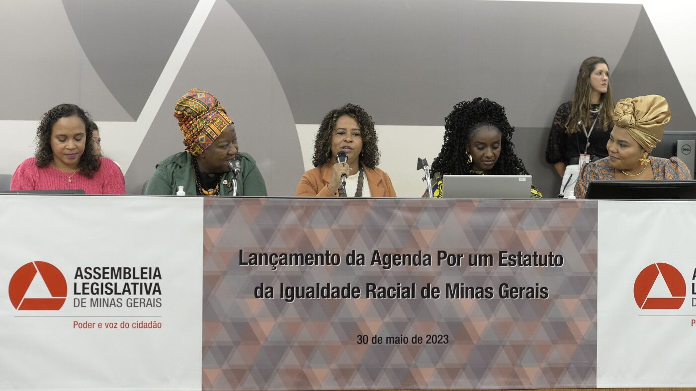 Fotografia colorida do lançamento da Agenda por um Estatuto da Igualdade Racial de Minas Gerais. Na foto, as deputadas que estão guiando o processo estão na bancada de destaque do auditório. Deputada Leninha está falando ao microfone.  