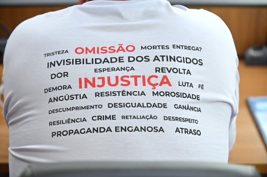 Parte de trás de uma camisa branca usada por um dos participantes da reunião. Contém as palavras: tristeza; mortes; entrega?; invisibilidade dos atingidos; dor; esperança; revolta; demora; angústia; resistência; e várias outras me preto. As palavras Omissão e Justiça estão maiores e em vermelho. 